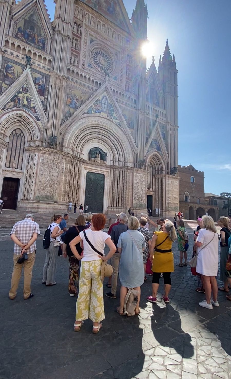 A group of people looking up at the banded, mosaic-covered facade of Orvieto Cathedral in the sun